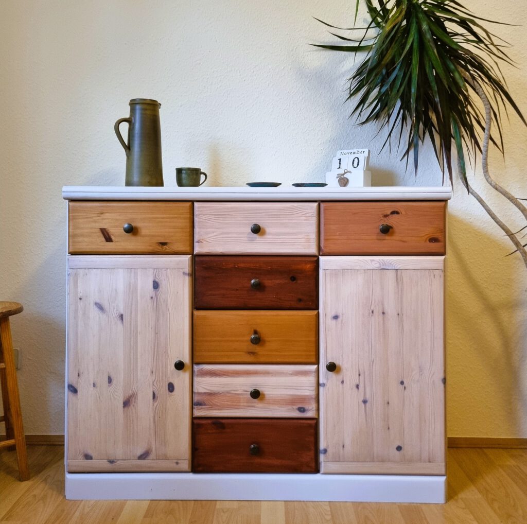 Rustikale Kommode aus DIY Upcycling Produktion der Firma TREWIENS aus Massivholz mit Schubladen in verschiedenen Holzfarben, dekoriert mit einer grünen Keramikkanne, einer Tasse und einem Kalender auf weißer Oberfläche, daneben eine große Zimmerpflanze.